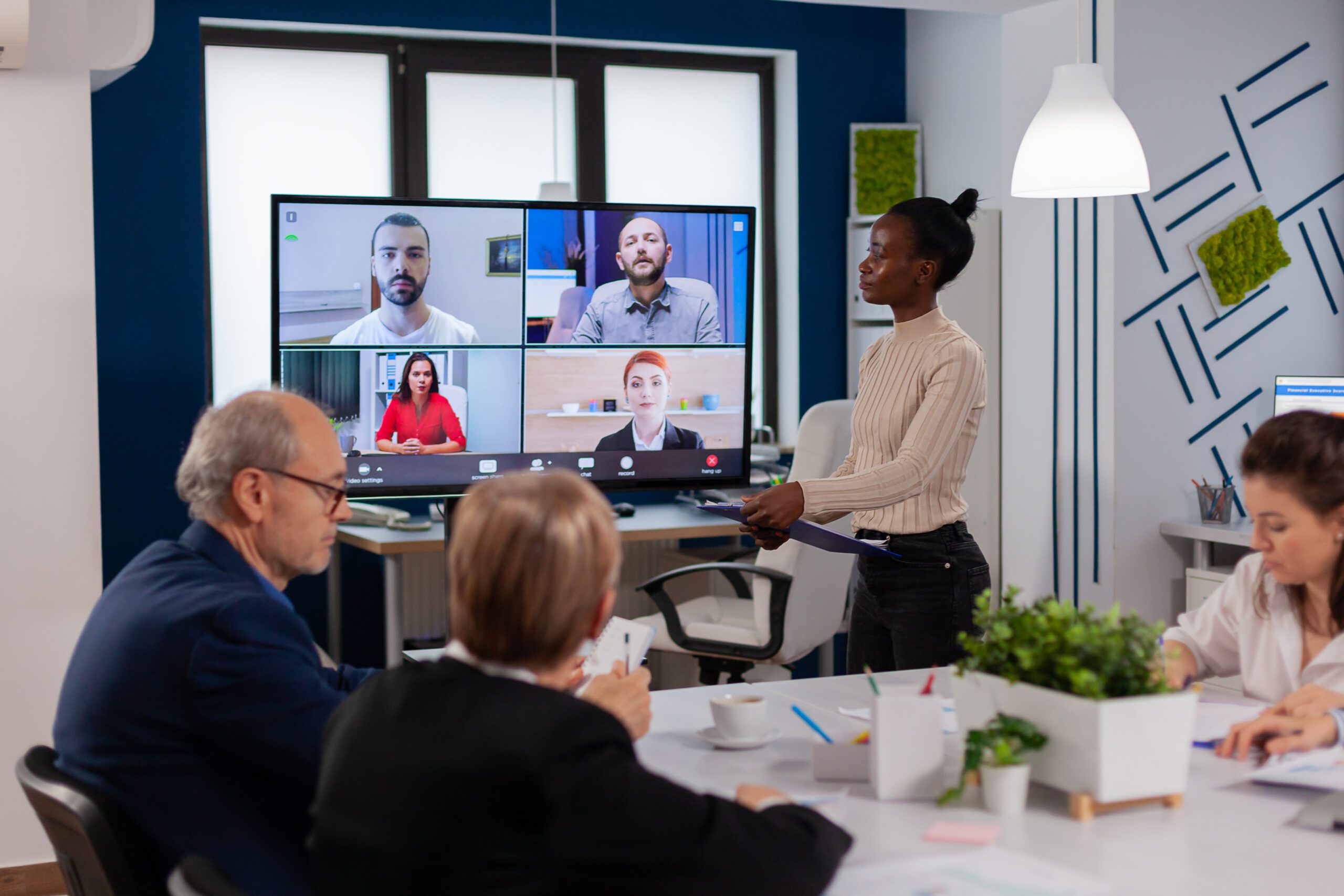 A woman enhances collaboration as she presents to colleagues in a meeting room, while a video conference call with four participants is displayed on a screen.