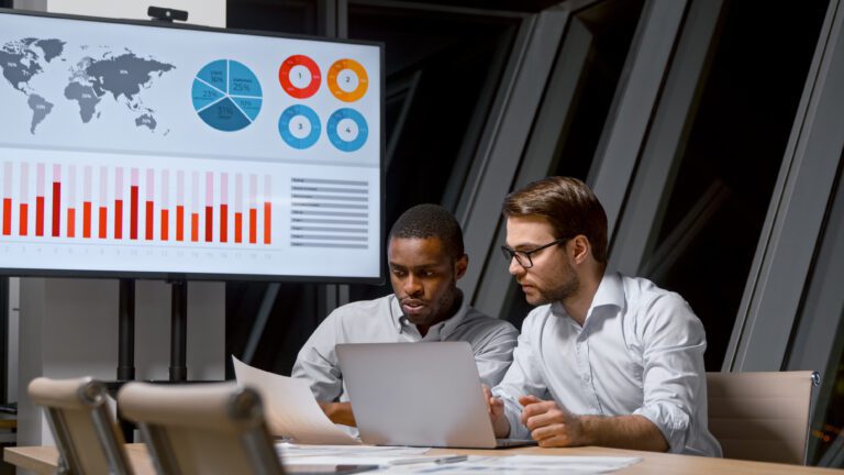 In a modern office setting, two men review documents and data on a laptop. A screen in the background displays a world map, pie charts, and bar graphs, highlighting tools that drive decisions with precision.