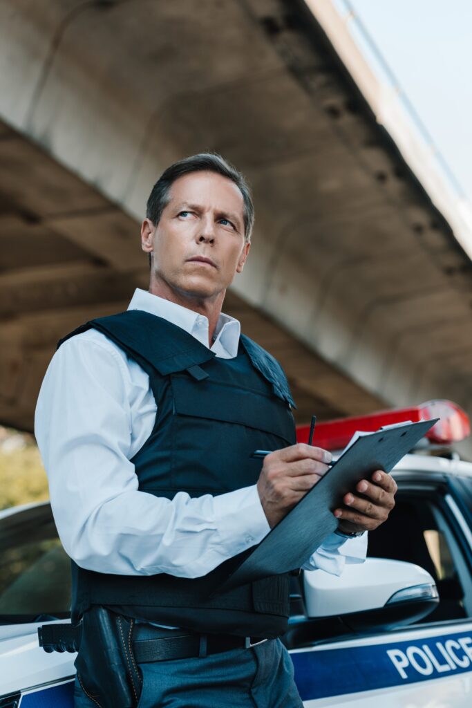 low angle view of middle aged male police officer in bulletproof vest writing in clipboard near car