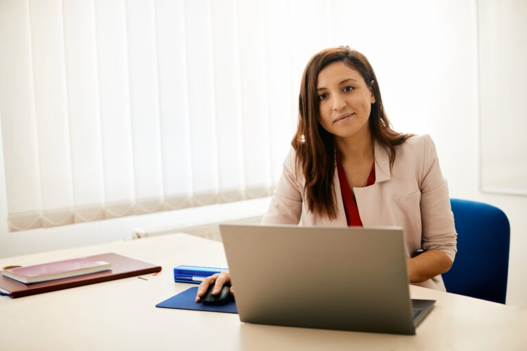 Smiling female teacher using laptop in the classroom and looking at camera.