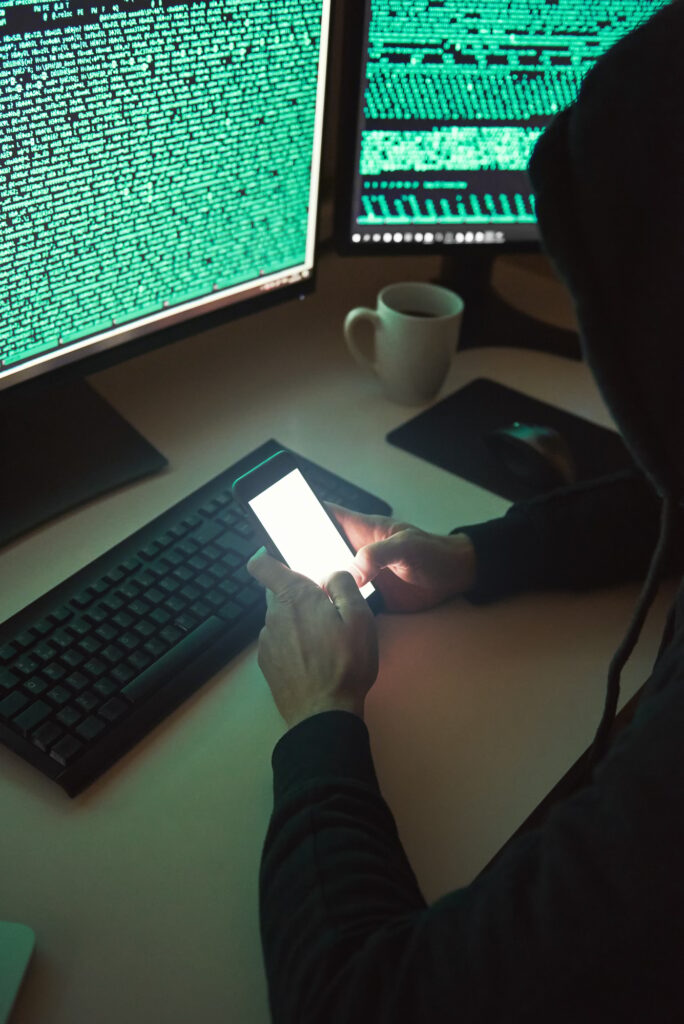 Vertical photo of young hacker in black hoodie using multiple computers