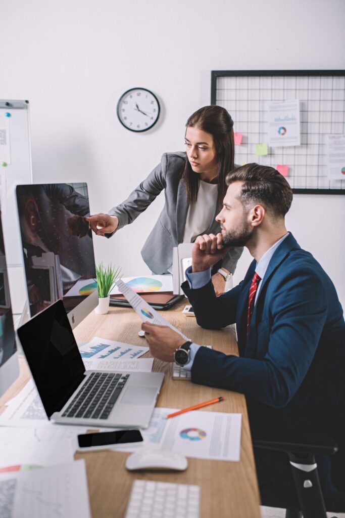 Data analyst pointing at computer monitor near colleague holding paper with charts at table